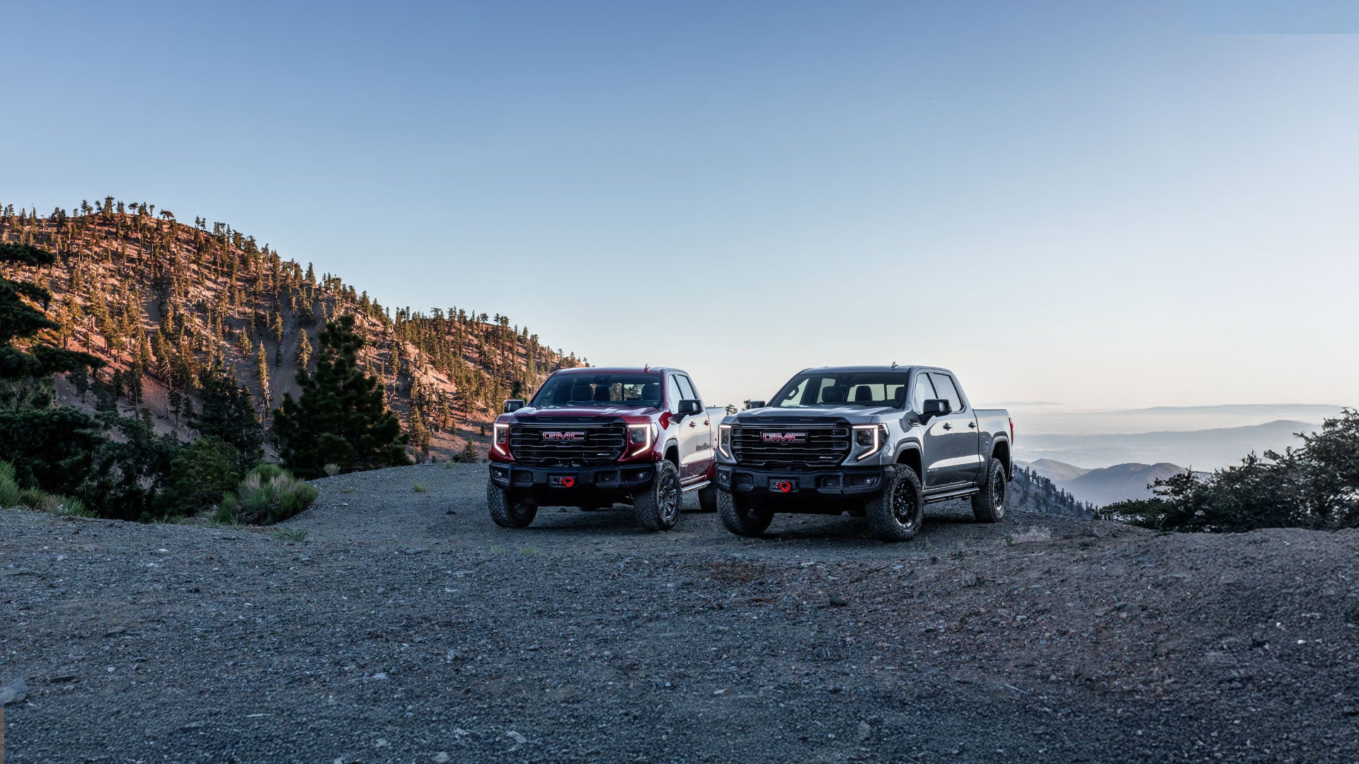 Two gray GMC Sierra trucks, one AT4X and one Denali, parked on a dirt road with mountains in the background.